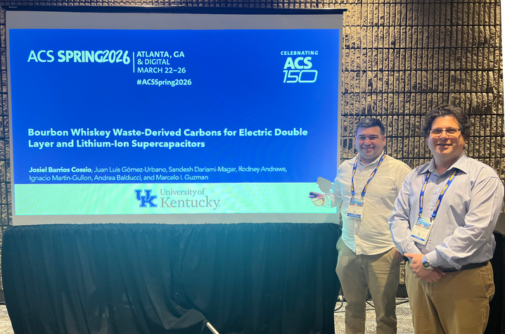 Photo of a conference poster on a screen titled “Bourbon Whiskey Waste‑Derived Carbons for Electric Double Layer and Lithium‑Ion Supercapacitors” at ACS Spring 2026 in Atlanta. Two attendees with badges stand beside the screen.