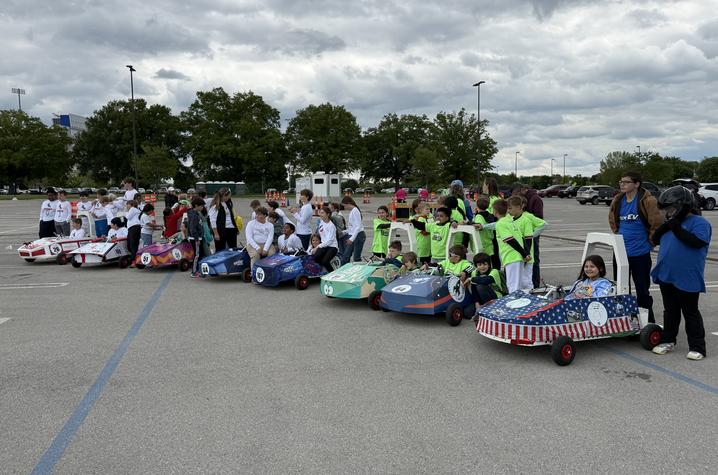 The competition field. Elementary student teams line up handmade electric race cars at the starting area of the GEN‑EV competition, with rows of students, teachers and spectators gathered in a large parking lot.