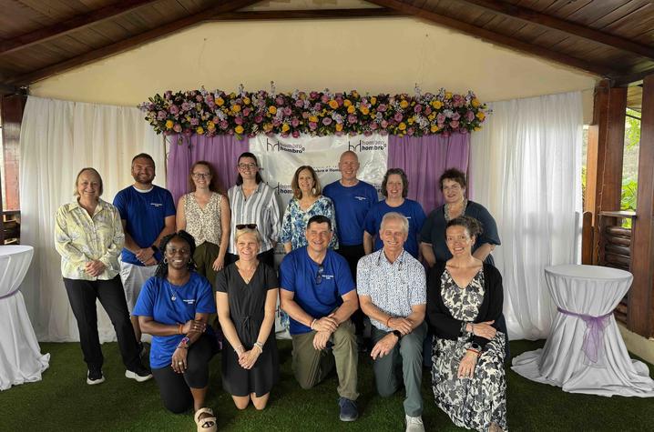 Group of about 14 adults pose for a photo under a wooden pavilion, arranged in two rows on green turf with white drapes and a colorful floral display behind them.