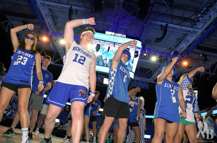 Photo of DanceBlue 2024 A group of college-aged dancers wearing blue athletic outfits perform in an arena under bright lights, raising their arms in unison. Some wear jerseys and shorts, and one dancer has “LEX 12” on their shirt.
