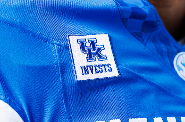 photo of UK Invests patch Close-up of a blue football jersey featuring a white patch with the words “UK INVESTS” and the University of Kentucky logo stitched near the shoulder.