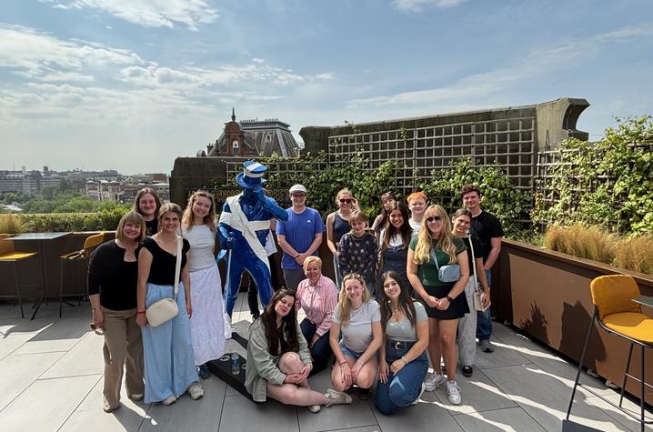 A group of students poses for a photo on a rooftop terrace alongside a person in a blue-and-white mascot costume.
