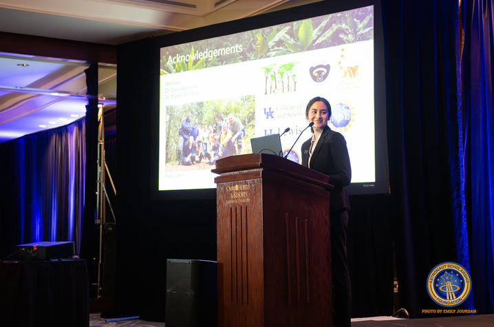 A person stands at a wooden podium presenting in a conference room. A large screen behind them displays a slide with photos of fieldwork and several logos. Blue curtains frame the stage, and an emblem is visible on the lower right side of the image.