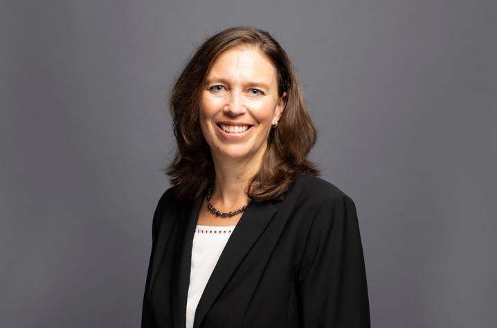 Jasinski posed for photo against a dark gray background wearing a black blazer and white blouse.