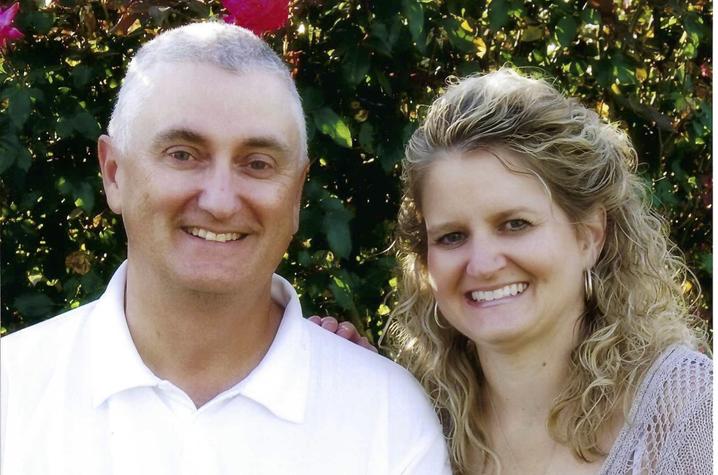 A smiling middle aged-white couple sits in front of a wall of flowering shrubbery. He has short gray hair and is wearing a white polo shirt with a UK logo. She has curly blond hair past her shoulders, wearing a loosely-woven lilac sweater. 