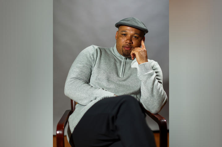 Black man wearing a grey sweater and black pants seated in a chair.