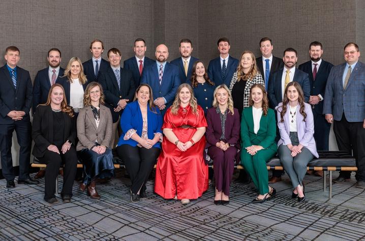 A group of adults posed for a formal photo in two rows inside a conference room.