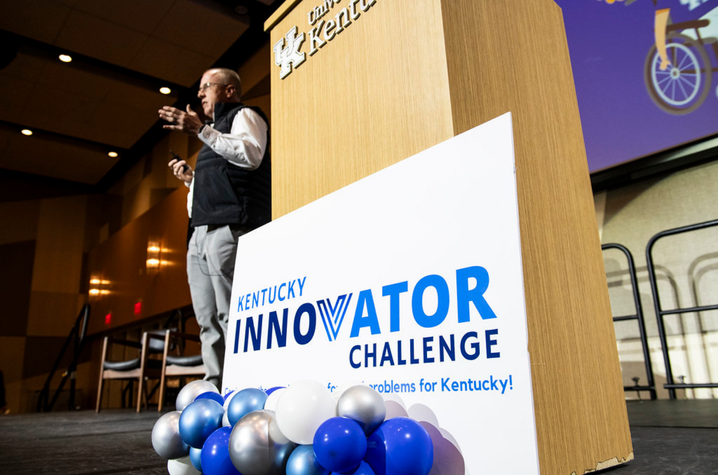 Kentucky Innovator Challenge sign Presenter speaks beside a University of Kentucky podium at the Kentucky Innovator Challenge. Blue, white and silver balloons decorate the stage in a large conference hall.