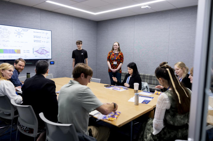 Several participants sitting at conference table in smaller room with two individuals standing, giving presentation.