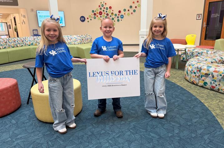 Three children wearing matching blue T‑shirts stand in a colorful lobby area, holding a large white sign that reads “Lexus for the Little Ones.” The space includes patterned seating, round ottomans, and bright decor on the walls.