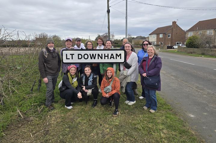 Group of people posing outdoors around a road sign reading “LT DOWNHAM.” The group stands and kneels beside a road, with hedges to the left and brick houses to the right. Overhead utility poles and wires run behind the sign under a cloudy, overcast sky.