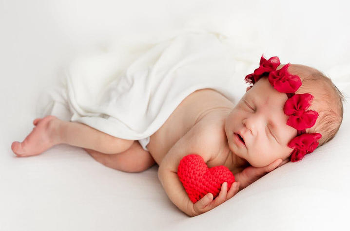 Image of Millie Wiseman with stuffed heart Newborn baby sleeps on a white blanket while holding a small red heart and wearing red ribbon bows on the head.