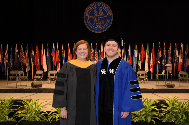 Macauley and Constantino Two people in University of Kentucky academic regalia stand on a commencement stage beneath the university seal. International flags line the background during a doctoral graduation ceremony.