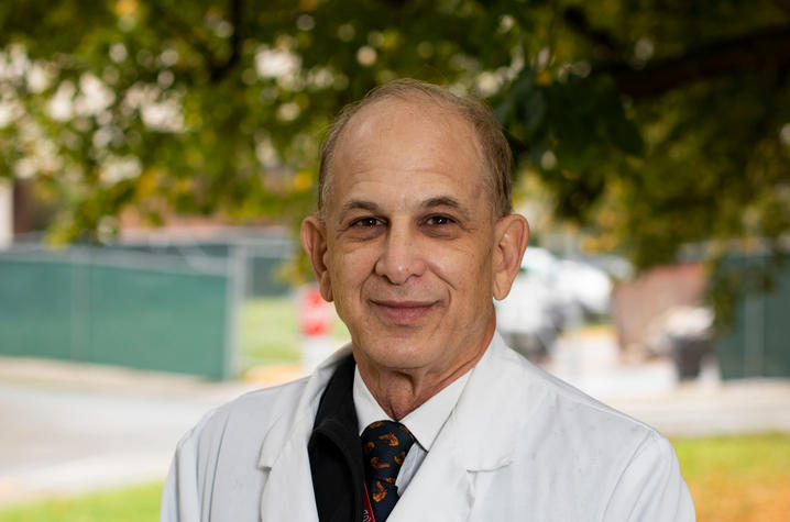 Dr. Richard Greenberg stands outside wearing his white coat. There is a tree behind him and colorful fallen leaves on the ground. 