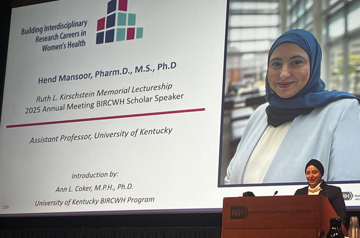 Photo of Hend Mansoor A presenter stands at a podium delivering a talk in front of a large projected slide. The slide shows a portrait and text about a research lecture on women’s health, including the speaker’s name, academic credentials, and affiliation with UK.