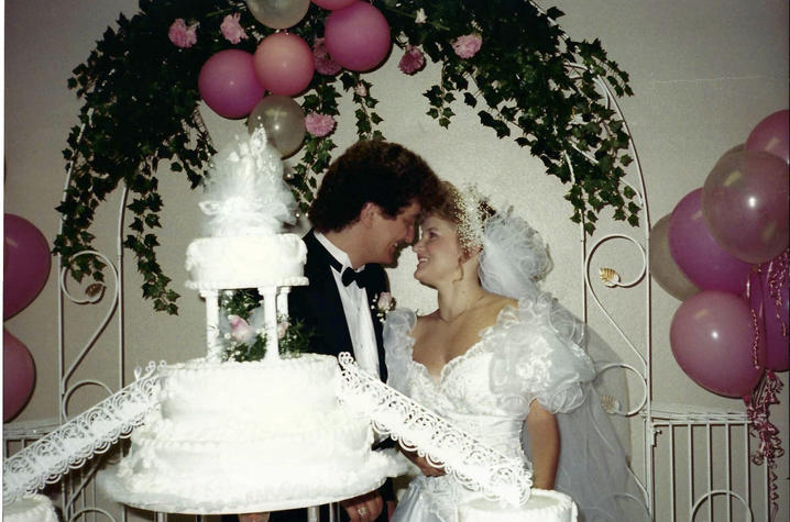 Photo of a young white couple at their wedding. A multi-tiered cake is in the foreground; the couple stand behind it, facing each other with foreheads touching and big smiles on their faces. They're framed by a canopy of foliage and pink balloons. 