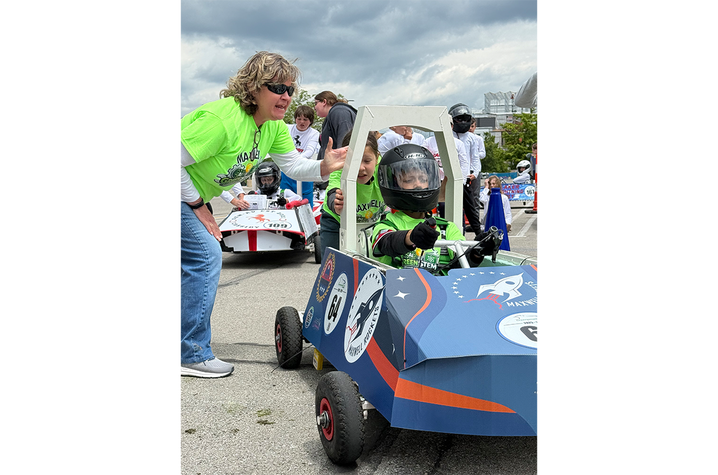 Go go go! An adult mentor leans toward a helmeted student seated in a handmade electric race car, giving instructions before the race while other teams wait nearby.