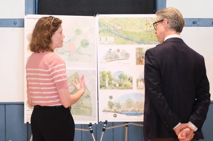 Two people stand in a gallery space reviewing design boards on easels, with one gesturing toward illustrated plans and renderings.