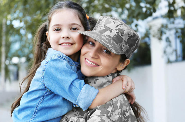 Photo of military member and child Person in military camouflage uniform embraces a young child outdoors near a building and trees.