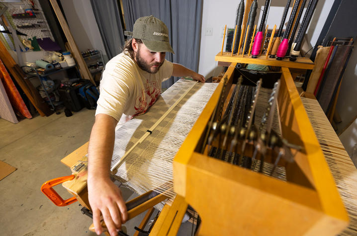 White male in a white t-shirt wearing a ball cap, seated at a loom. 