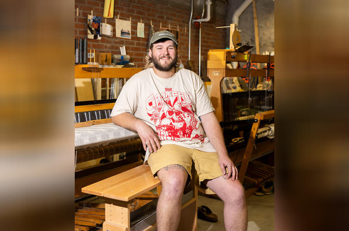 White male in a white t-shirt with a red graphic on the front, also sporting a ball cap, seated at a loom looking at camera.