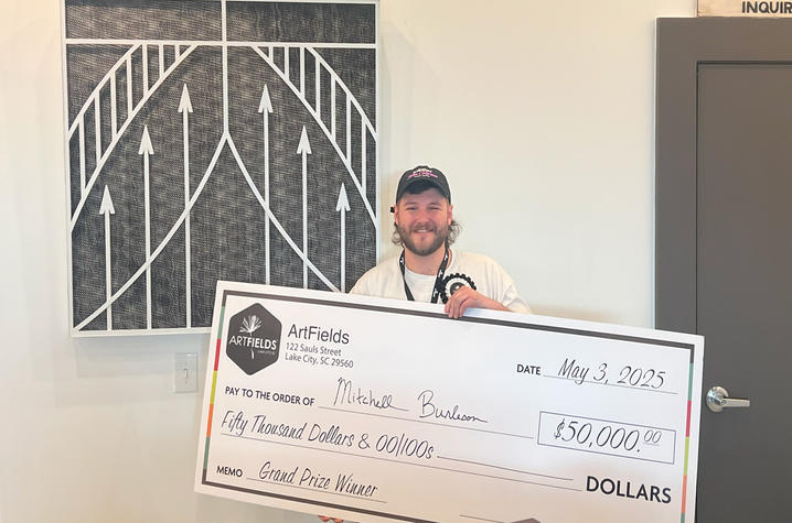White male in white t-shirt and ball cap, holding a large-sized ceremonial check, standing in front of a fiber art piece.