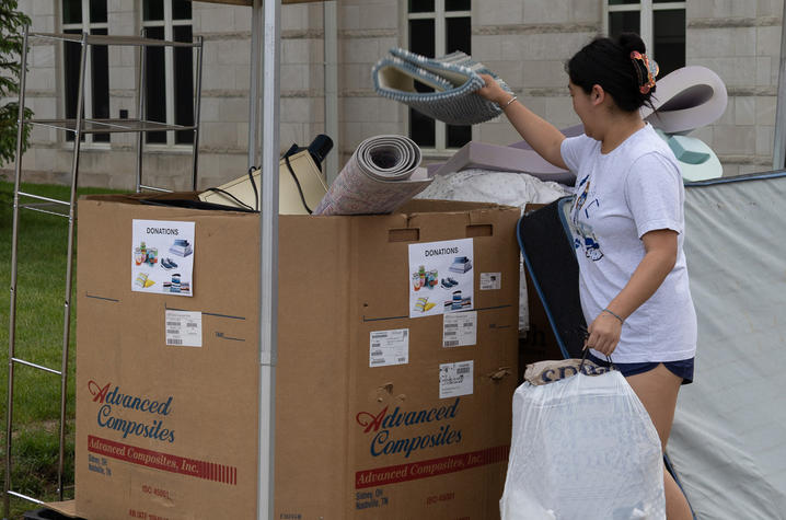 A person places items into a large cardboard collection box labeled for recycling outside a building.