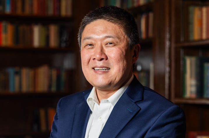 A person in a navy suit and white shirt stands with arms crossed in front of bookshelves filled with books.