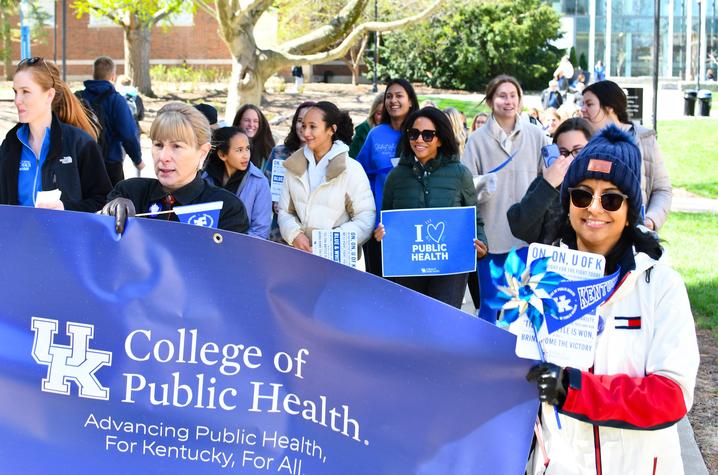 Group marching outdoors holding a “UK College of Public Health” banner and signs during a campus parade.