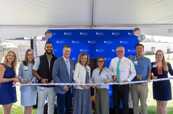 Image of ribbon cutting at Centerpoint Health – Paris ribbon cutting Nine people participate in a ribbon-cutting ceremony under a white tent, standing in front of a blue UK Golisano Children’s Network backdrop and holding gold ceremonial scissors as they cut a white ribbon.