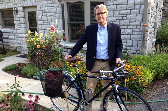 Ned Crankshaw wearing a blazer and blue button down shirt standing on the sidewalk with a bicycle in front of a stone house. 