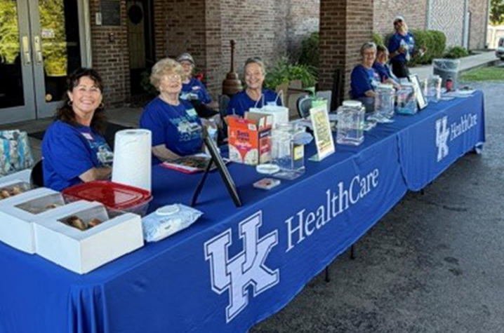 Photo of Markey OCS team at community event Several people sit behind tables covered with blue UK HealthCare tablecloths at an outdoor community event, with informational materials, containers and refreshments displayed on the tables.