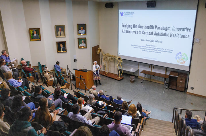 One Health Symposium presenters Presenter stands at the front of a lecture hall, speaking to an audience seated in tiered rows. A large projection screen displays a presentation titled “Bridging the One Health Paradigm.”