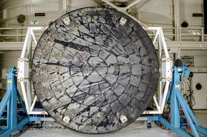 Large circular spacecraft heat shield mounted on blue supports inside an industrial facility.