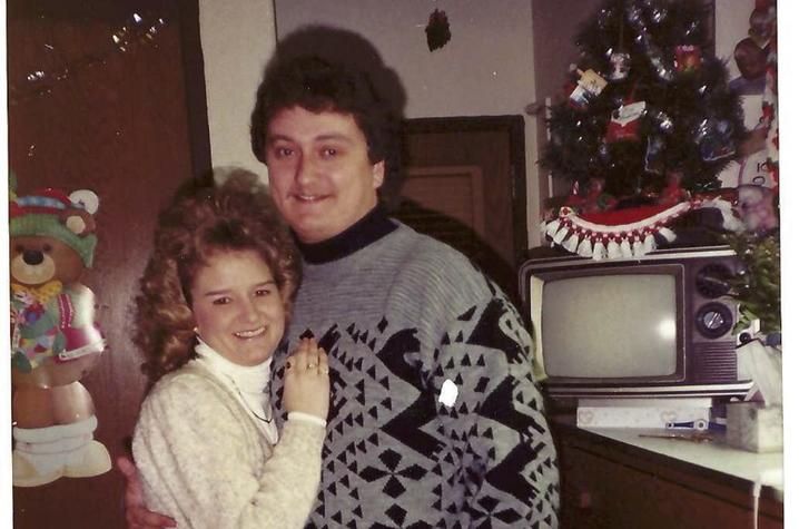 Photo from the late 80s of a young couple in a dorm room with Christmas decor. She's petite, with curly blond hair, wearing a cream sweater and red pants. He's tall, with dark wavy hair, wearing a black and gray patterned sweater with black trousers. 