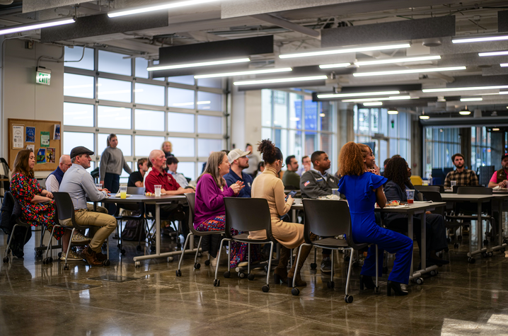 A group of people sit at tables in a large, modern, open indoor space with bright overhead lighting and polished concrete floors. 