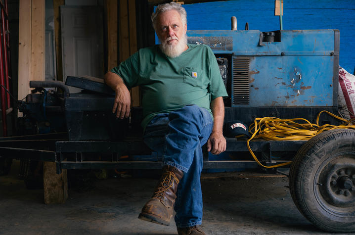 Person in a green shirt and work boots sitting on a trailer with industrial equipment and coiled yellow extension cord in a workshop.