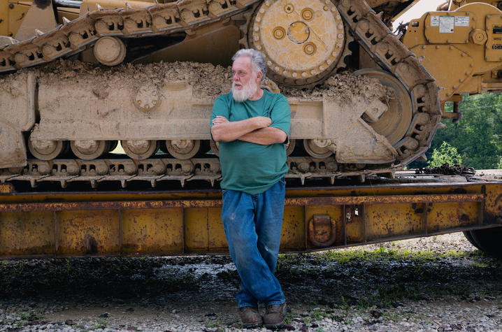 Person in a green shirt and jeans leaning against a large, muddy bulldozer track outdoors.