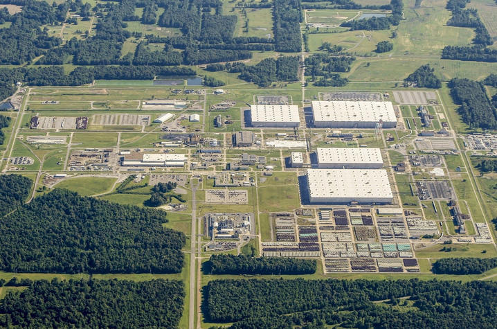 Aerial view of a large industrial complex surrounded by dense forest and open fields. The facility includes multiple large white-roofed warehouse-style buildings, grid-like roadways, smaller structures, parking areas, and neatly arranged outdoor storage.