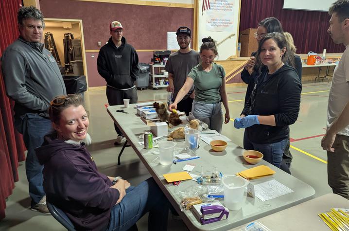 A group gathers around a table covered with stuffed animals, containers, tools and other supplies during a hands-on activity in a large multipurpose room.