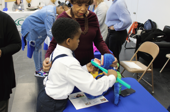A child practices brushing a large model of teeth with guidance from an adult at a health education table.