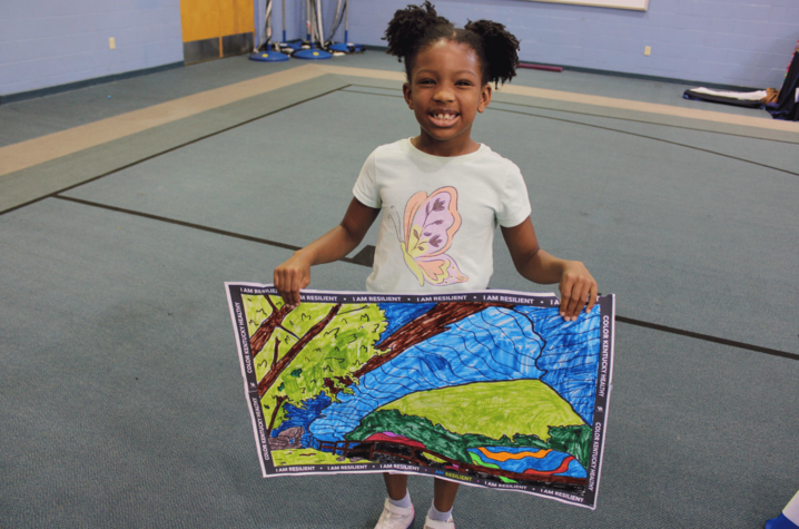 A child holds up a colorful nature drawing labeled “I Am Resilient” at a community health event.