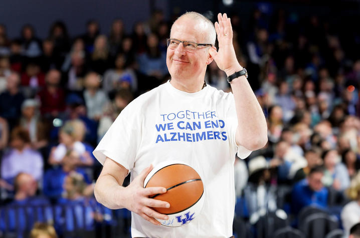 A man wearing a white T-shirt that reads “Together We Can End Alzheimer’s” smiles while holding a University of Kentucky basketball on a court. A crowd of spectators sits blurred in the background, suggesting a game-day or special event setting.