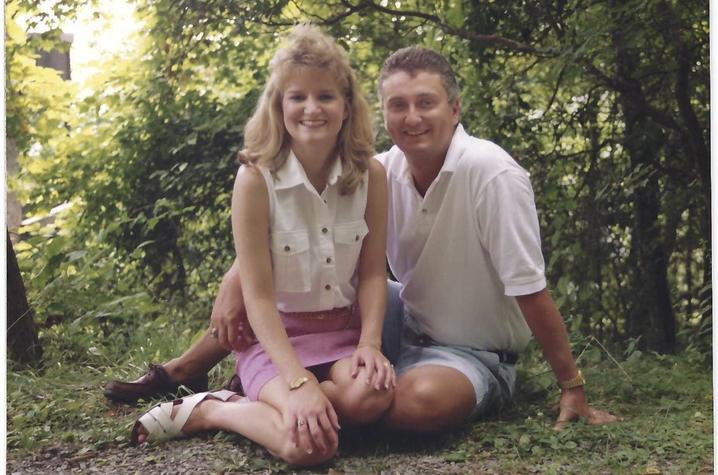 A young white couple sit in a wooded area, smiling at the camera, She has blond shoulder-length hair and wears a sleeveless white blouse, thigh-length pink shirt, and white sandals. He's wearing a white polo, nice jean shorts, and brown loafers. 