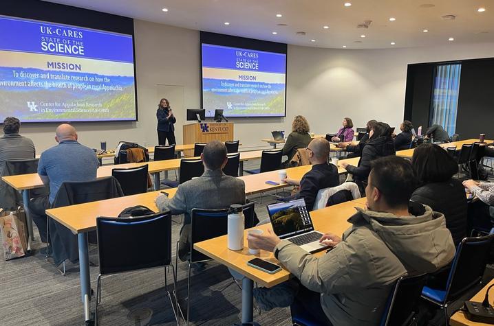 A group of attendees sits at tables in a conference room listening to a speaker presenting at the front. Two large screens display a “UK-CARES State of the Science” slide about environmental health research in rural Appalachia. People take notes on laptop