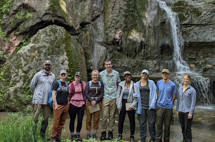 Photo of 2026 Potter summer interns and KGS mentors Nine people stand in front of a rocky waterfall and moss-covered cliff in a forested area.