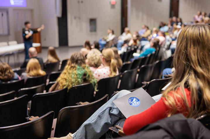 A lecture hall filled with people seated in rows of black chairs, facing a presenter at the front of the room. The presenter stands near a projection screen and gestures while speaking.