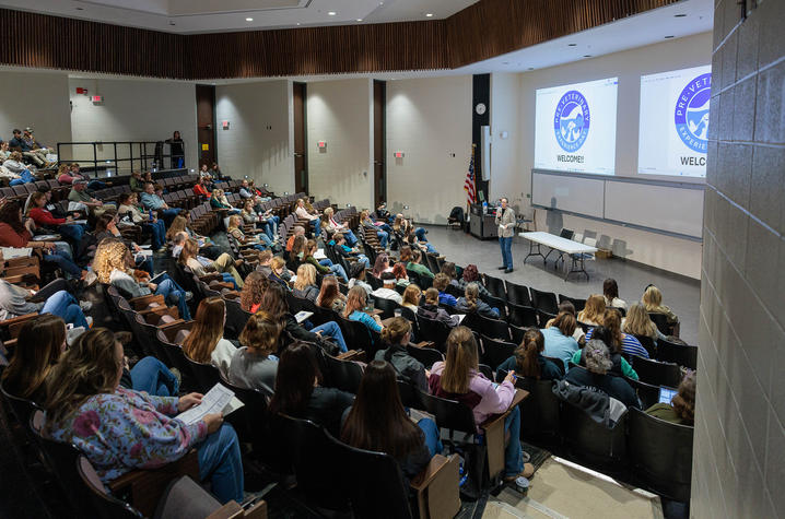 A large lecture hall filled with many attendees seated in tiered rows, facing a presenter at the front of the room. The presenter stands near a podium and gestures toward two large projection screens displaying circular blue-and-white logos. 