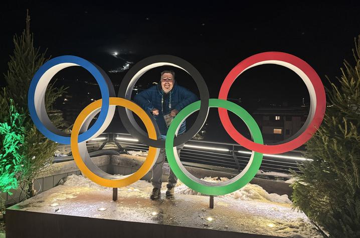 A man stands inside the Olympic rings display at night with snowy mountains in the background.