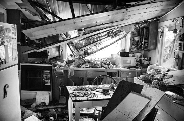 Terry’s childhood home Black-and-white photograph of a kitchen severely damaged by a storm. The roof has collapsed inward, exposing beams and debris across counters, tables and appliances, illustrating the destructive impact of a natural disaster.
